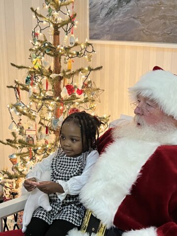 a little girl sits on Santa's lap with a lighted Christmas tree behind them.