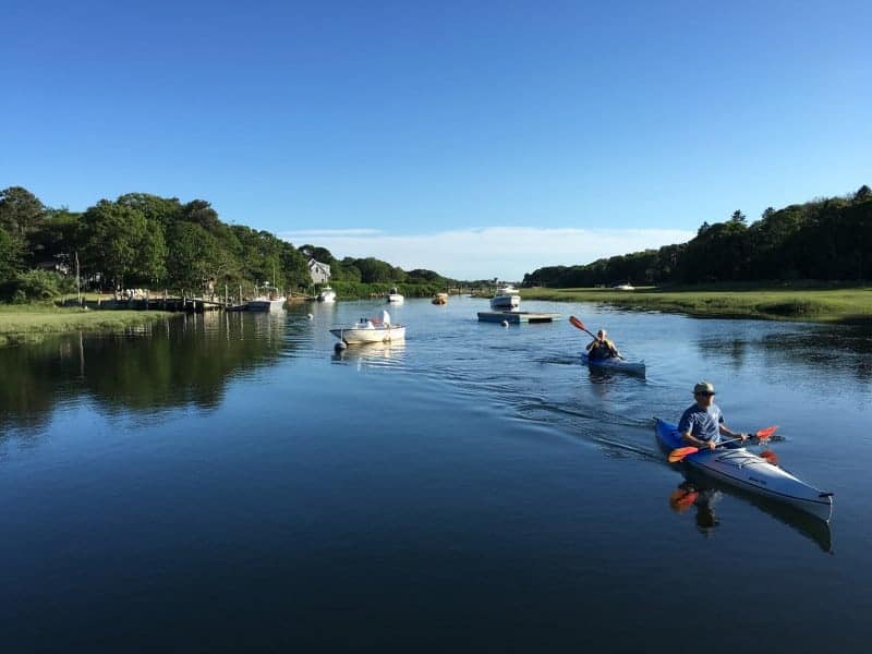 Two people in kayaks paddle their way down a river where there are several boats moored and the shores are green with marshes and trees