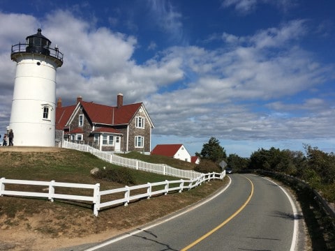 Photo of Nobska Lighthouse in Woods Hole. A white lighthouse beside a shingled house with a red roof. A white picket fence along the yard.