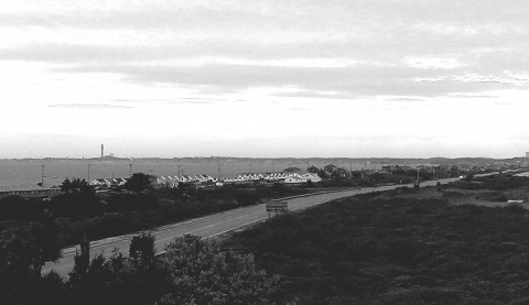 A black and white photo showing a highway in the foreground and then some small beach cottages and the Pilgrim Monument towering in the sky in the background.