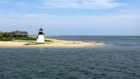 White lighthouse on the sandy shore of Nantucket Island, surrounded by deep blue water and a light blue sky