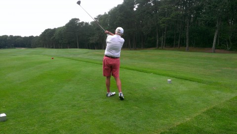 A man golfing.  His club is in the air. He is wearing red shorts, a white polo shire and a white cap.  The grass is deep green and trees surround the golf course.