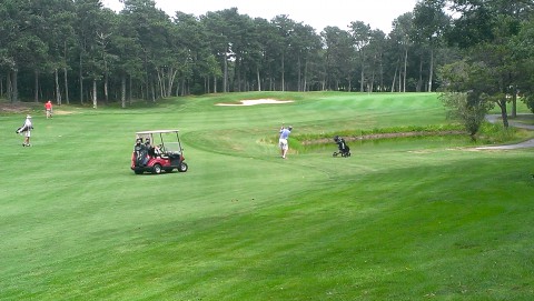 A green fairway on a golf course surround by trees with a sand trap in the near distance.  3 men are walking along the course and there is a golf cart in the middle.