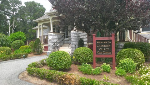 Exterior of the Cranberry Valley Golf course.  The entrance has landscaping with many shrubs, and the building is shingle style.  The entrance sign is framed in red.