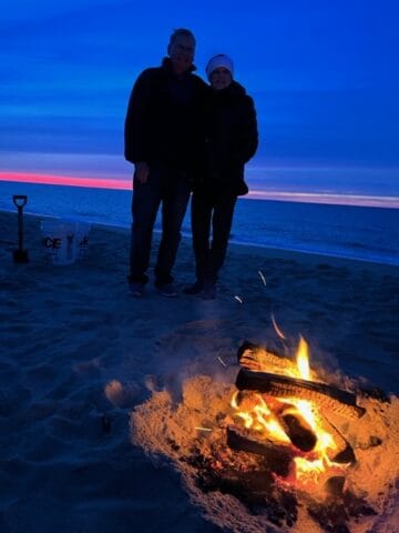 At dusk, two people in silouette on the beach with the sunset in the background and a fire in the foreground