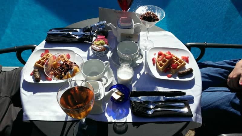 A table by a pool set for breakfast, including tea cups, waffles, glass of juice, yogurt parfait and napkinds and silverware