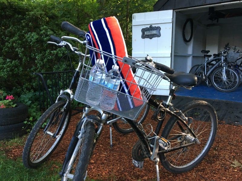 Two bicycles with water bottles and red, white and blue beach towels sit outside of a shed filled with more bicycles