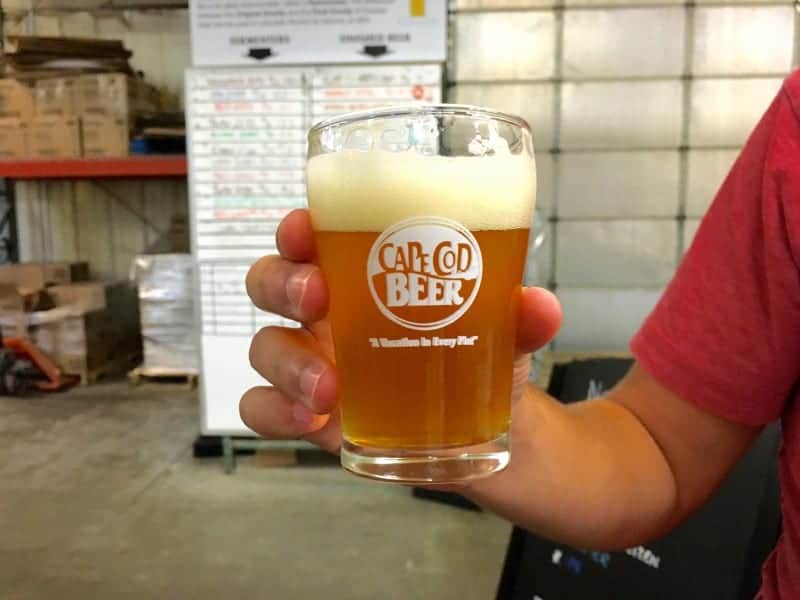 A person in a red shirt holding a glass of beer. picture taken inside a warehouse with palates of materials in the background.