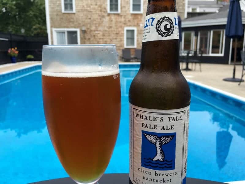 A glass of beer and beer bottle in front of a pool.  A singled building can be seen in the background.