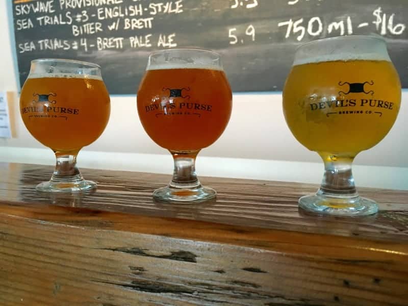 Wooden table with glasses of beer in different shades of amber.  Behind on the wall is  a black board listing the types of beer available.