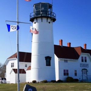 White lighthouse and building against a blue sky.  Several flags fly on a pole in front of the lighthouse