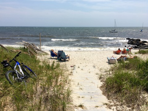 At the end of a sandy boardwalk is a beach and ocean. A bike is parked in the sea grass, people are relaxing on beach chairs and several sailboats with sails down, are in the ocean