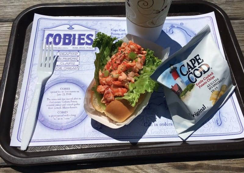 a lobster roll on a tray with a bag of potato chips a drink and a white fork.