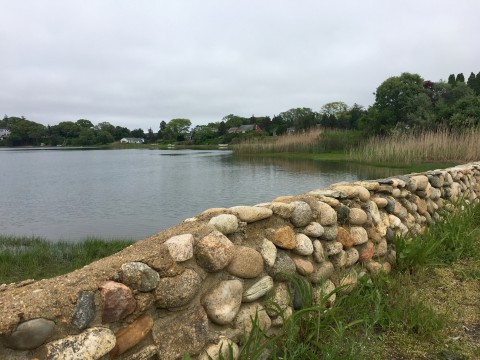 A cobblestone wall is in the front of a pond surrounded by grasses and trees