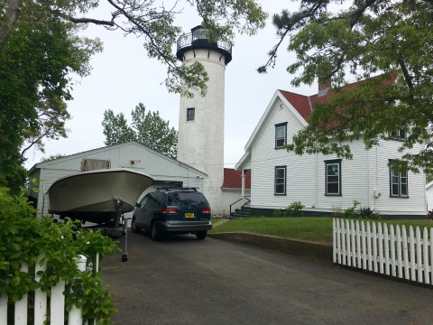 White lighthouse. A truck and boat are in the driveway. Lighthouse is next to a white building with a red roof