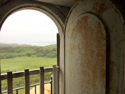 View from the inside of the lighthouse to outside through an arched window.  view is through green fields to the ocean