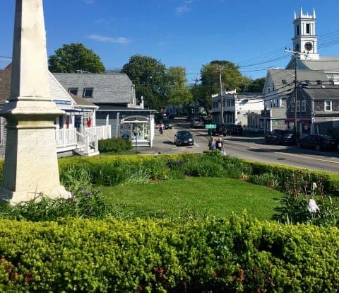 a white monument in a green town square with a street and white church in the background