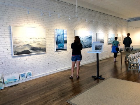 A girl looking at art hanging on a wall at a gallery.  The walls are white brick and the wooden floors have rugs