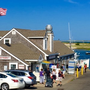 The building of the Chatham Pier Fish market with people walking by, parked cars and a view to the ocean and a boat