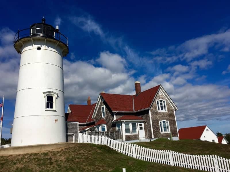 White lighthouse and the light housekeepers building with a red roof and shingled walls.  A white picket fence runs down a grassy area.  Deep blue sky with white wispy clouds