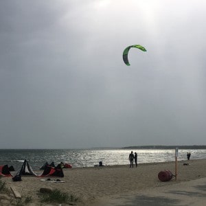 An overcast sky with a wind sail, kite in the sky, and people on a sandy beach by the ocean