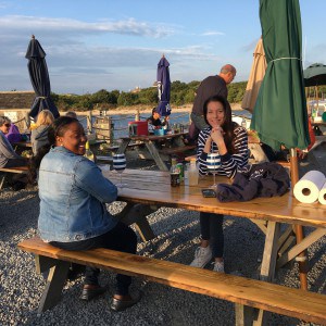 two ladies sitting at an outdoor seaside picnic table with a closed green umbrella