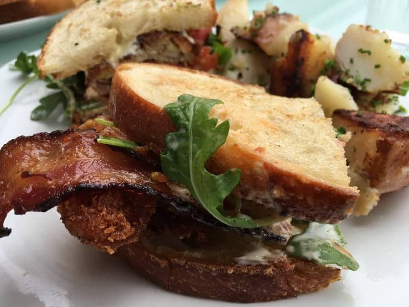 Close up of a sandwich with bacon and arugula, a side of potato salad