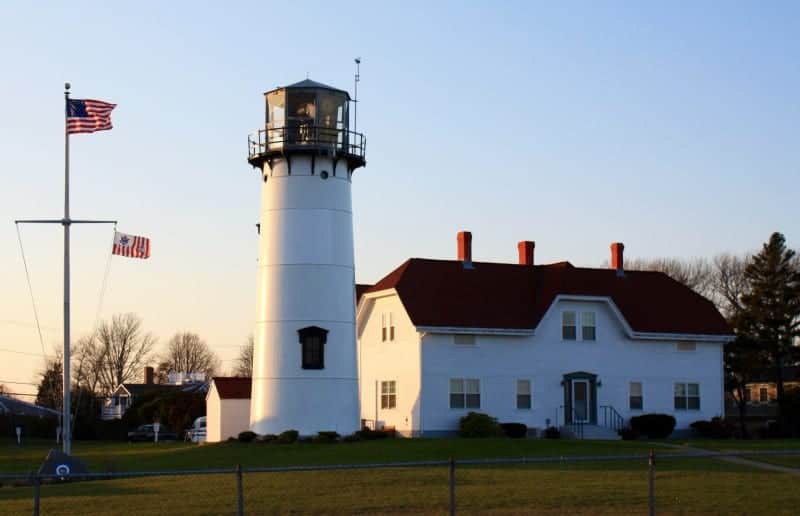 a light house in Chatham that is white and building behind is white with a red roof.  The American flag is flying on a flag pole