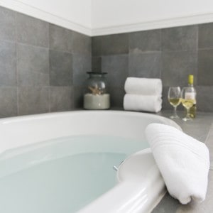 white bathtub surrounded by grey tiles. White towels, a bottle of wine and two glasses on the side.