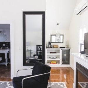 A room with a black chair, tv above a gas fireplace, a full length mirror, a dry bar with mini fridge and an open door leading to a bathroom.