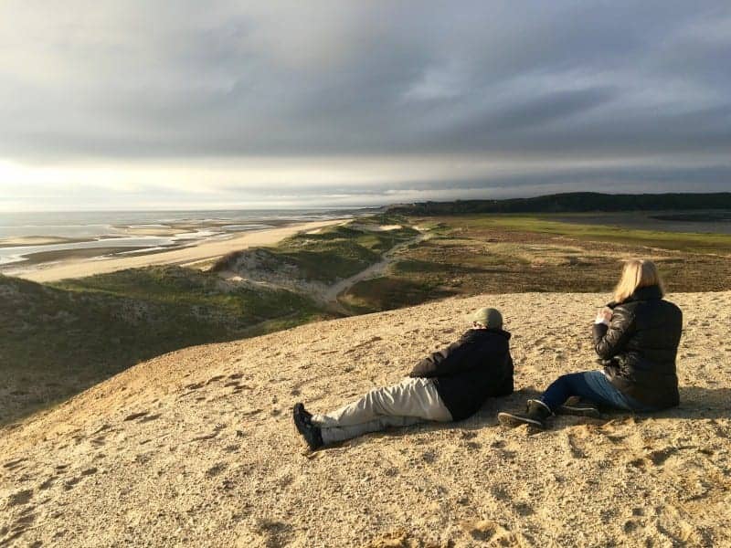 A couple sitting atop a dune looking toward the ocean on an overcast day