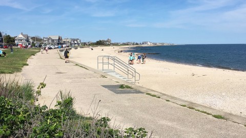 A beach in Falmouth with a cement boardwalk.  People are walking by and some houses are in the background