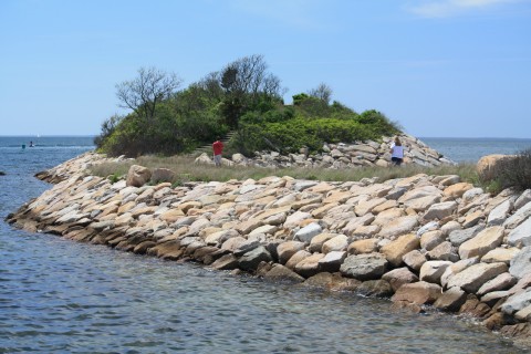 A rocky breakwall juts out into the ocean.  People are walking on top.  There is a lot of green vegetation