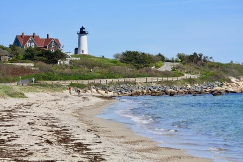 White Nobska Lighthouse is next to a brown building with a red roof.  A sandy beach and the ocean are in the foreground