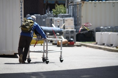 A man wearing a blue jacket and green knapsack is pushing a propeller on a white push cart