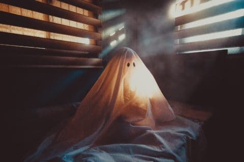 someone under a sheet that looks like a ghost with light streaming in from wooden slated windows
