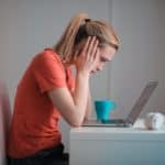 A women at a desk looking at a computer with her head in her hands. She is wearing an orange shirt and there is a teal coffee mug by her side and headphones
