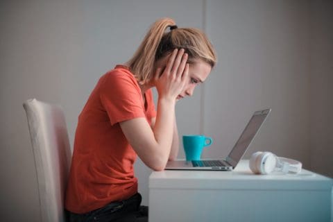 a woman at her desk doing remote work, staring pensively at a laptop while holding her head in her hands
