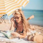 Girl in a bathing suit with sunglasses lying on a beach towel in the sand by the ocean reading a book and a yellow and white umbrella near by