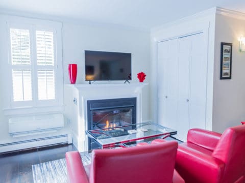 Bedroom with red leather chairs and a glass coffee table in front of a fireplace with a tv above it and red accent pieces. Double folding doors lead to a closet