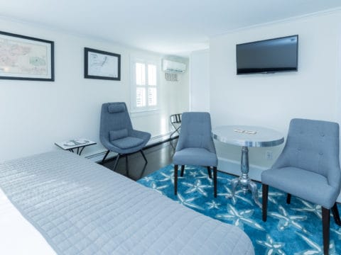 Bedroom sitting area. Walls are white, floor is dark brown with a blue rug with starfish. Silver table and 2 grey chairs are on the rub and a tv above the table. A reading chair to the side with small table