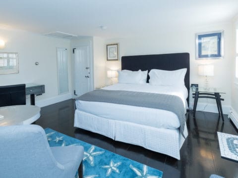 Bedroom with white bedding, grey bed saddle, black headboard, nights stands on either side with lamps and pictures above. A silver table aand 2 grey chairs sit on a blue rug with starfish. A mirror is on the wall and a desk with black leather chair
