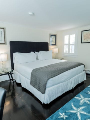 bedroom showing a bed with black headboard, white linens and grey bed saddle with glass nightstands beside the bed with crystal lamps and pictures above them. A blue rug with starfish. windows have shutters