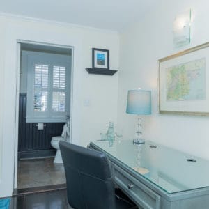 bedroom with a desk and black leather chair with a lamp and picture of a map and light above it. A shelf with a picutre of blue and black and open door leading to a bathroom