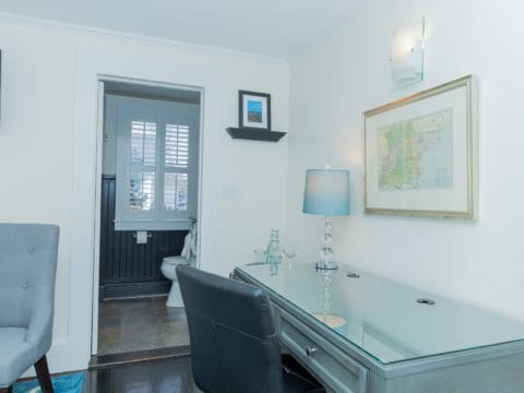 bedroom with a desk and black leather chair with a lamp and picture of a map and light above it. A shelf with a picutre of blue and black and open door leading to a bathroom