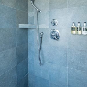 Inside of a shower with grey tiles, bath products on the wall and chrome faucets and hand held shower cord