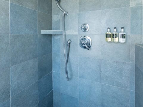 Inside of a shower with grey tiles, bath products on the wall and chrome faucets and hand held shower cord