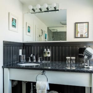 bathroom vanity with black granite top, black wainscoting, mirror with lights above it and 2 pictures on the wall.