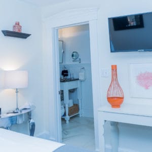 bedroom in white with orange accents, a tv on the wall and looking into the bathroom