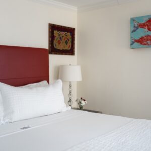 White bed with red leather headboard. A side table with a lamp, art on the wall